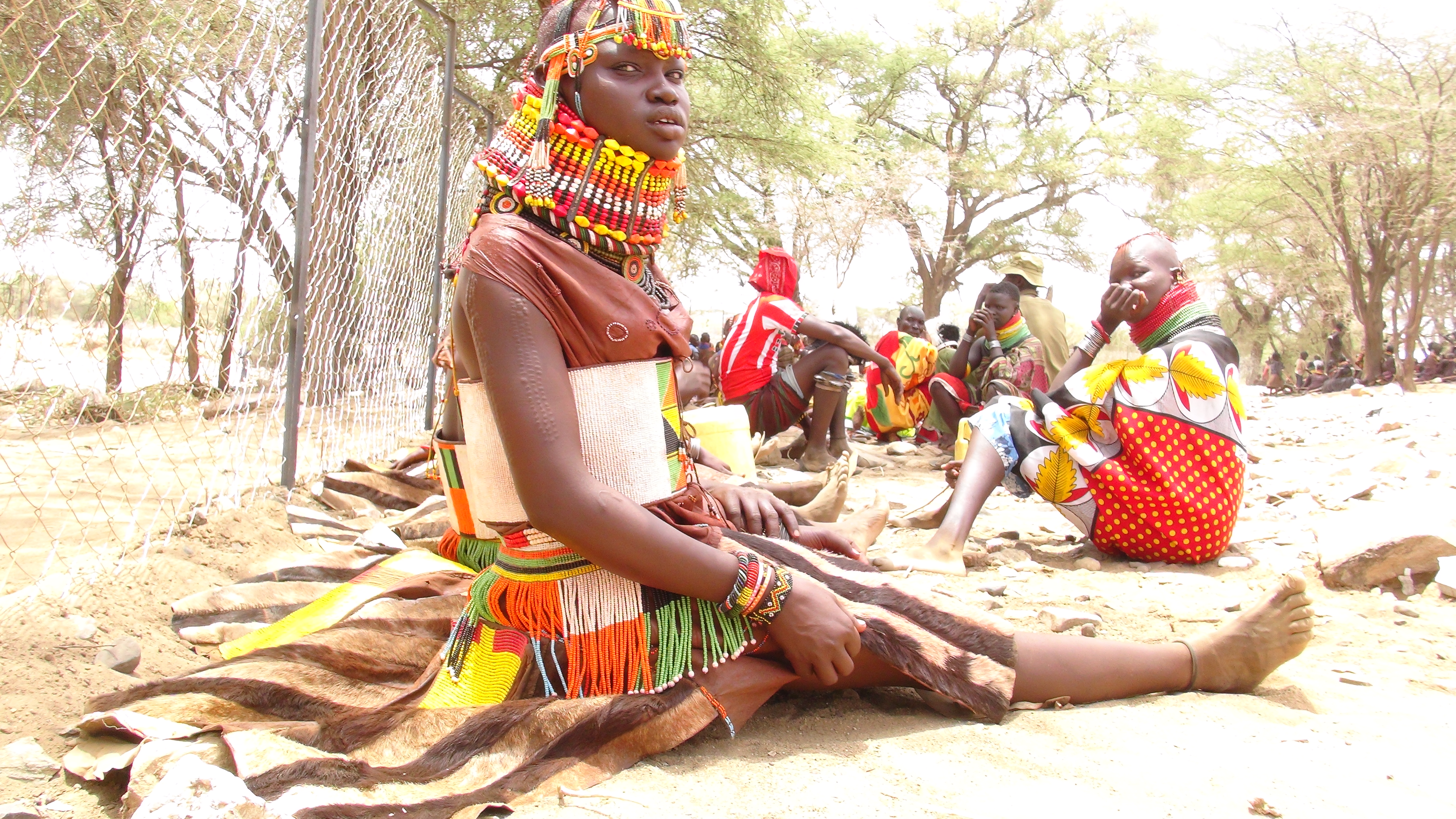 Women sit outdoors beneath trees, wearing colourful bead adornments and cloth or hide garments; the photographer identifies them as Turkana women attending a cross-border event in Uganda.