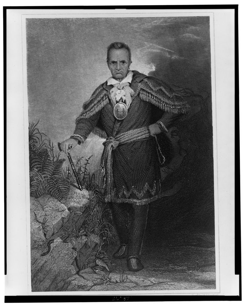 Historical engraved full-length portrait of Seneca leader Red Jacket, wearing a fringed coat and medal and holding a tomahawk beside rocks; black-and-white reproduction.