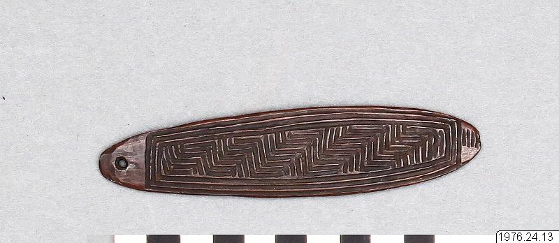 An elongated dark wooden object with a hole near one end and closely incised angular patterns, shown complete with a scale.