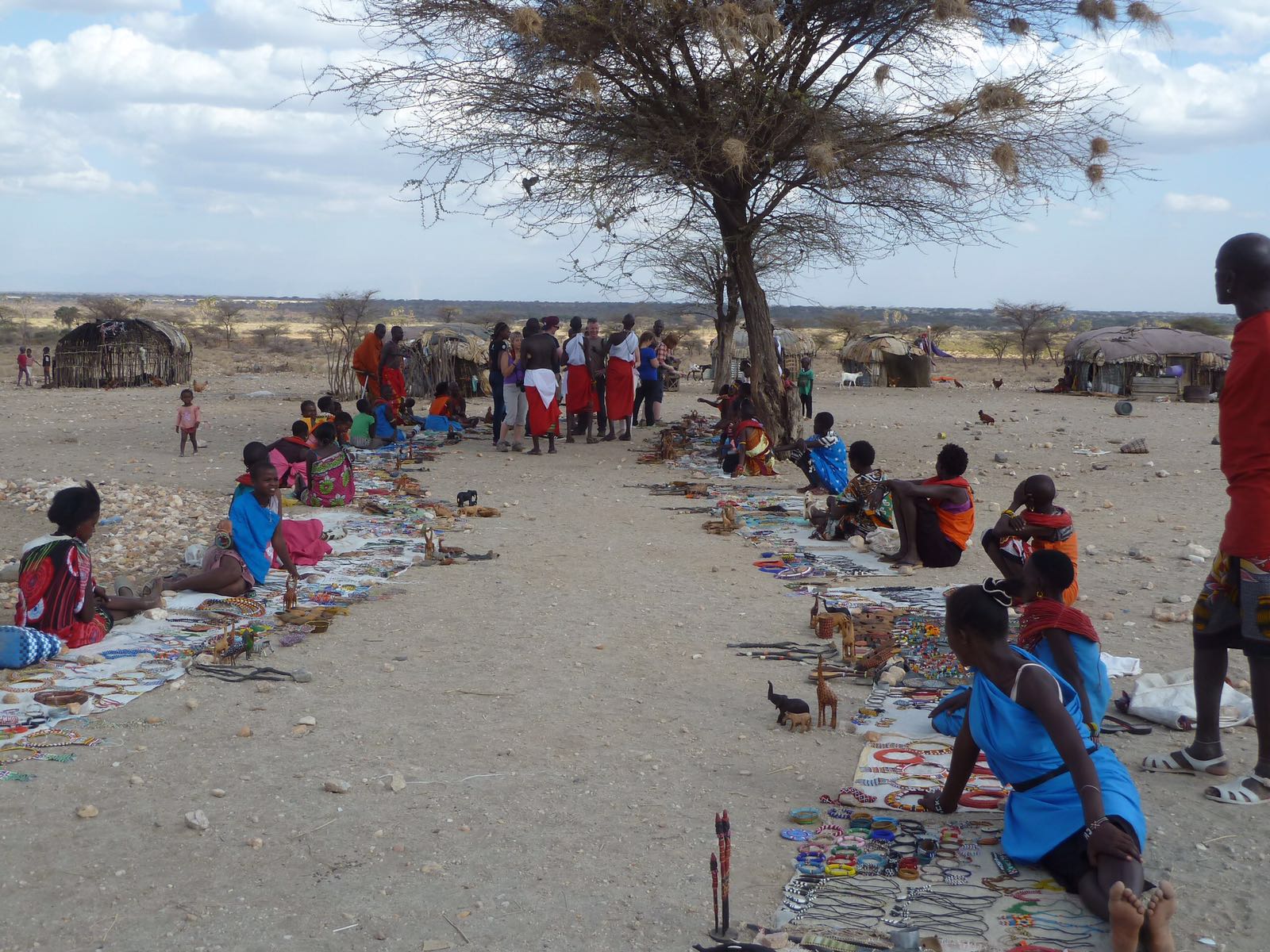 People sit beside rows of crafts on cloths beneath a tree, with several visitors and rounded buildings behind them; the photographer identifies the craft sellers as Samburu women.