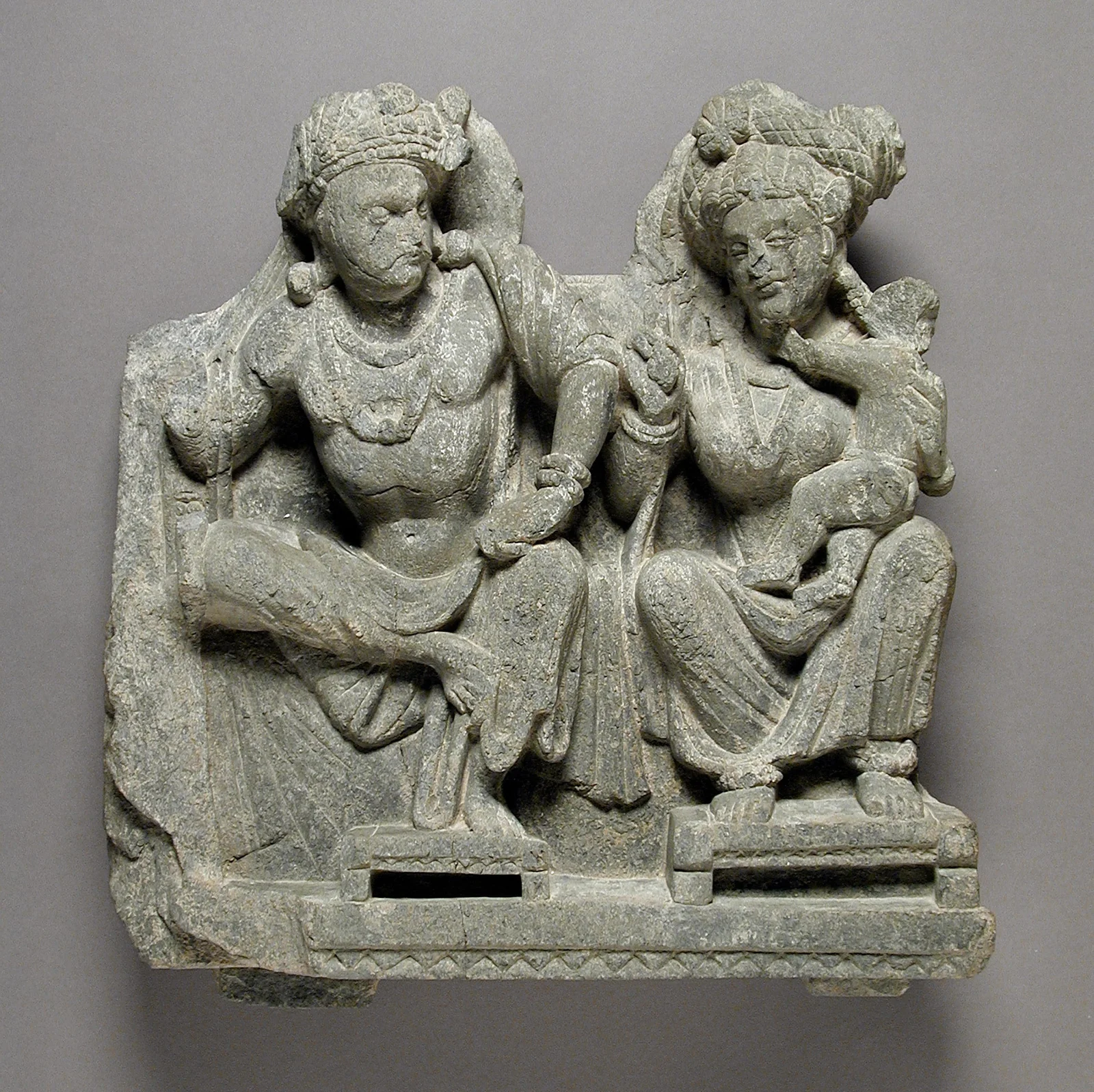 Grey schist relief of seated Panchika at left, wearing a turban and moustache, and Hariti at right with a child embracing her neck; both have halos, draped garments and individual footstools.