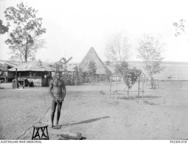 Black-and-white photograph of an unnamed man standing near a low stool, trees and settlement buildings, including a tall pitched roof.