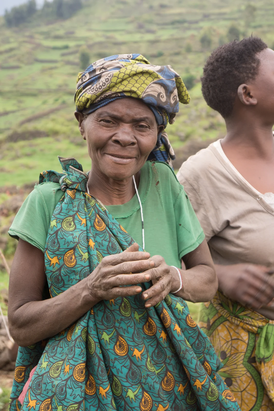 Woman identified by the photographer as Batwa in Kisoro, Uganda, wearing a green shirt and patterned headwrap and holding her hands together, with another person partly visible beside her.