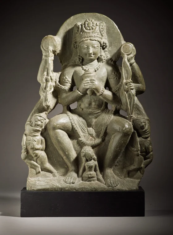 Grey stone sculpture of crowned Vishnu seated with several arms, holding the two small defeated figures of Madhu and Kaitabha at either side; the whole carved slab and black display base are visible.