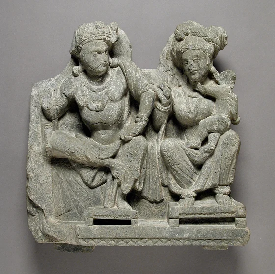 Grey schist relief of seated Panchika at left, wearing a turban and moustache, and Hariti at right with a child embracing her neck; both have halos, draped garments and individual footstools.