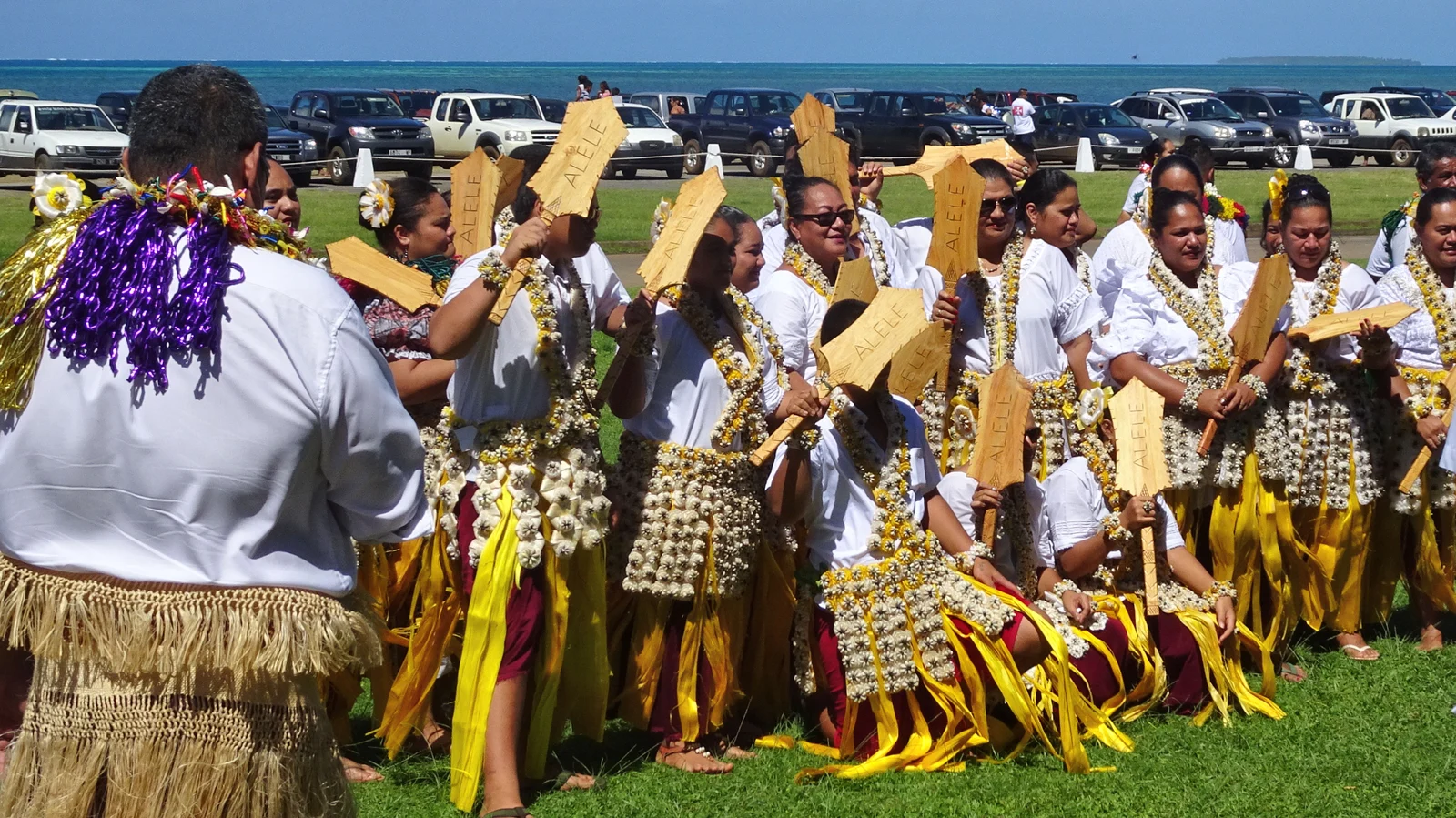 A group in white shirts, shell-covered adornments and yellow skirts holds woven fans marked ALELE beside a waterfront during the Wallis territorial festival.