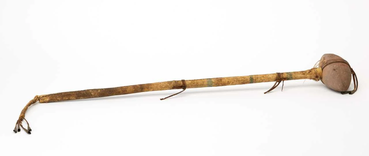 A complete long-handled stone-headed club with leather ties and small dangling metal bells on a white background.