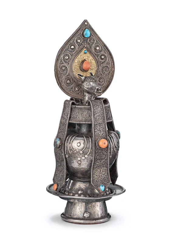 Silver-and-gold filigree ritual vase with a projecting horse head beneath a tall teardrop-shaped finial, inset with turquoise and coral; the complete vessel and pedestal are visible.