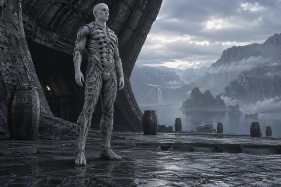 Pale humanoid in a ribbed suit overlooks rocky waters.