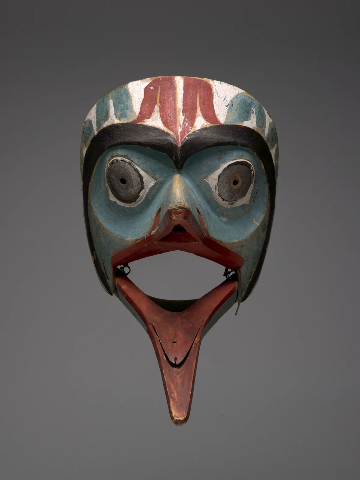 Front view of a carved bird mask with blue eye fields, black brows, red and white forehead designs, round eye holes and a long hinged red lower beak.