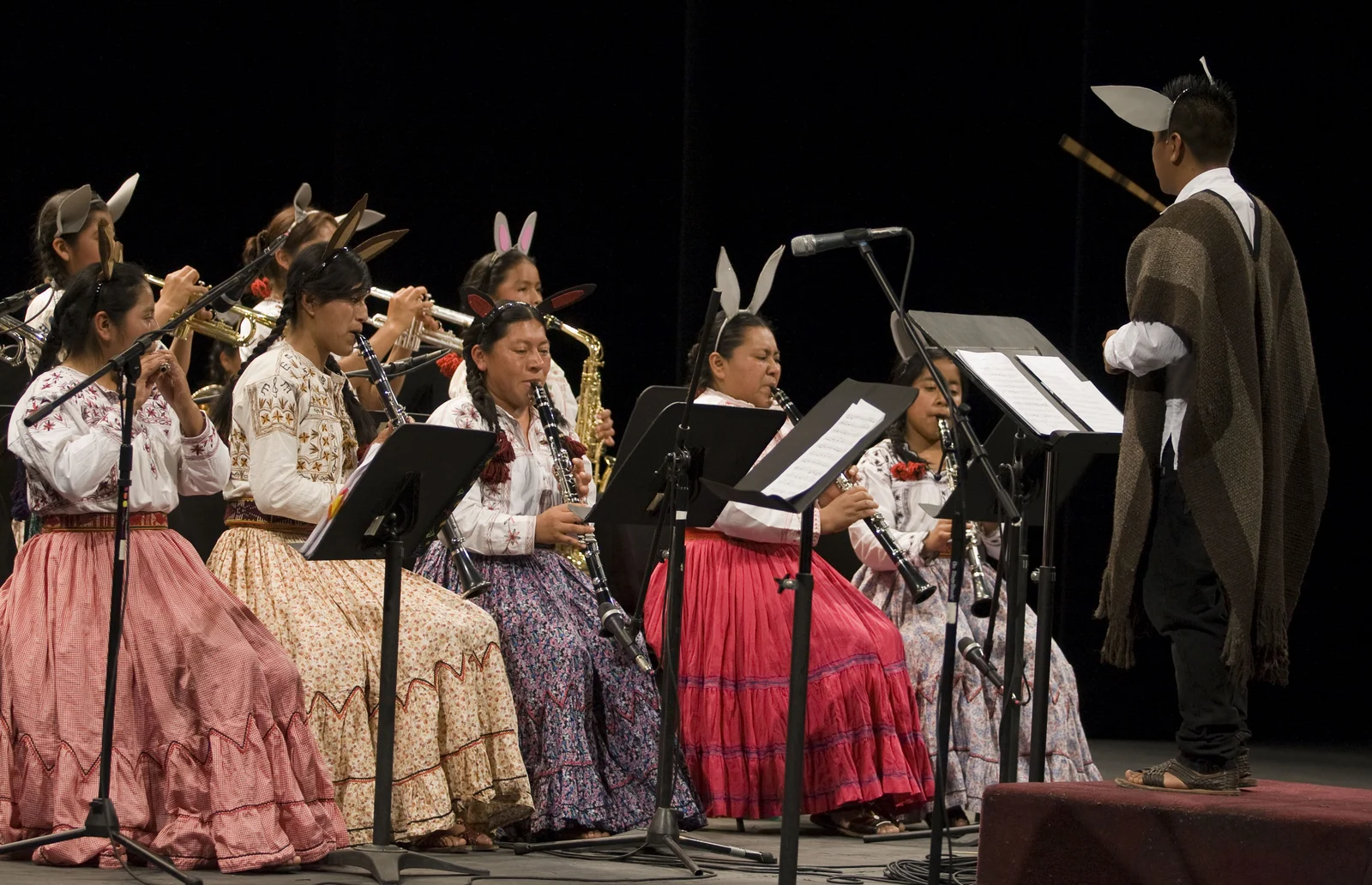 Musicians of Mujeres del Viento Florido play clarinets, flute and brass on stage while a conductor faces them; several wear theatrical animal-ear headbands.