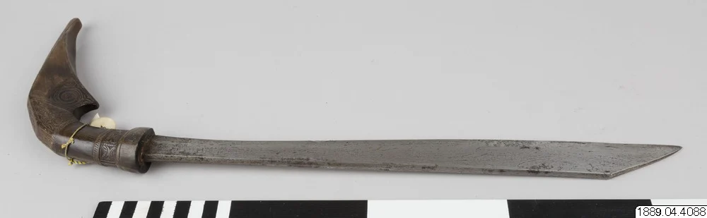A complete sword with a long metal blade and a strongly curved, carved dark hilt, shown above a scale and inventory label.