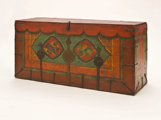 Red-brown Tibetan wooden trunk with two confronting green-bodied, golden-maned lions in red lobed cartouches; patterned borders and three pierced metal straps cross the painted front.