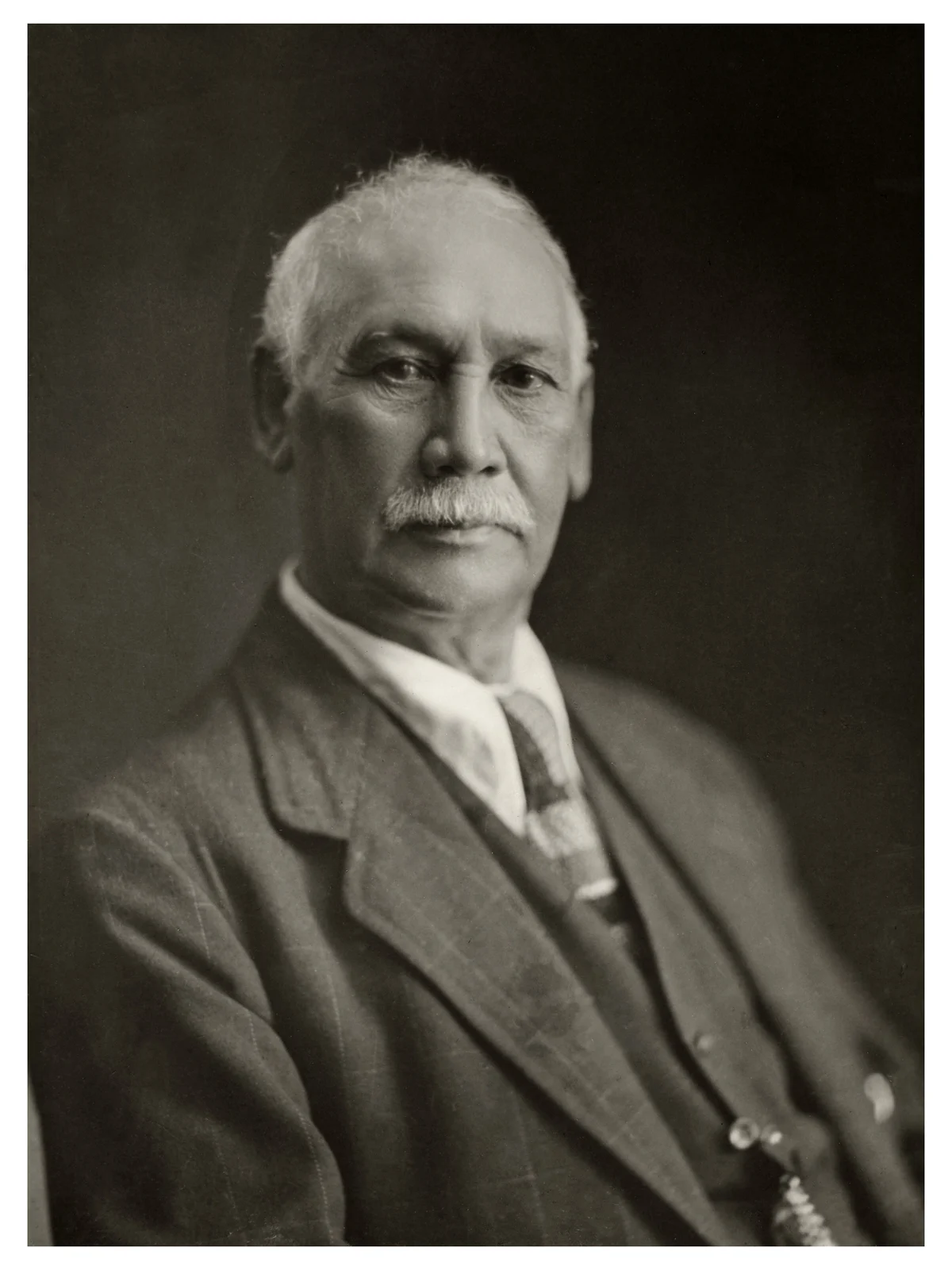 Monochrome seated portrait of Sir James Carroll wearing a dark suit, white shirt and patterned tie, with complete photograph borders visible.