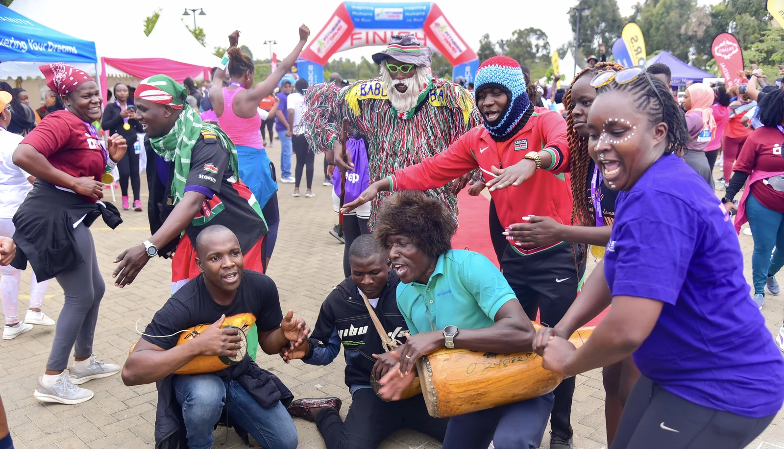 Two men crouch while playing cylindrical wooden drums beside a cheering runner and other performers at a public running-event finish line; the photographer identifies the musicians as Isukuti drummers.