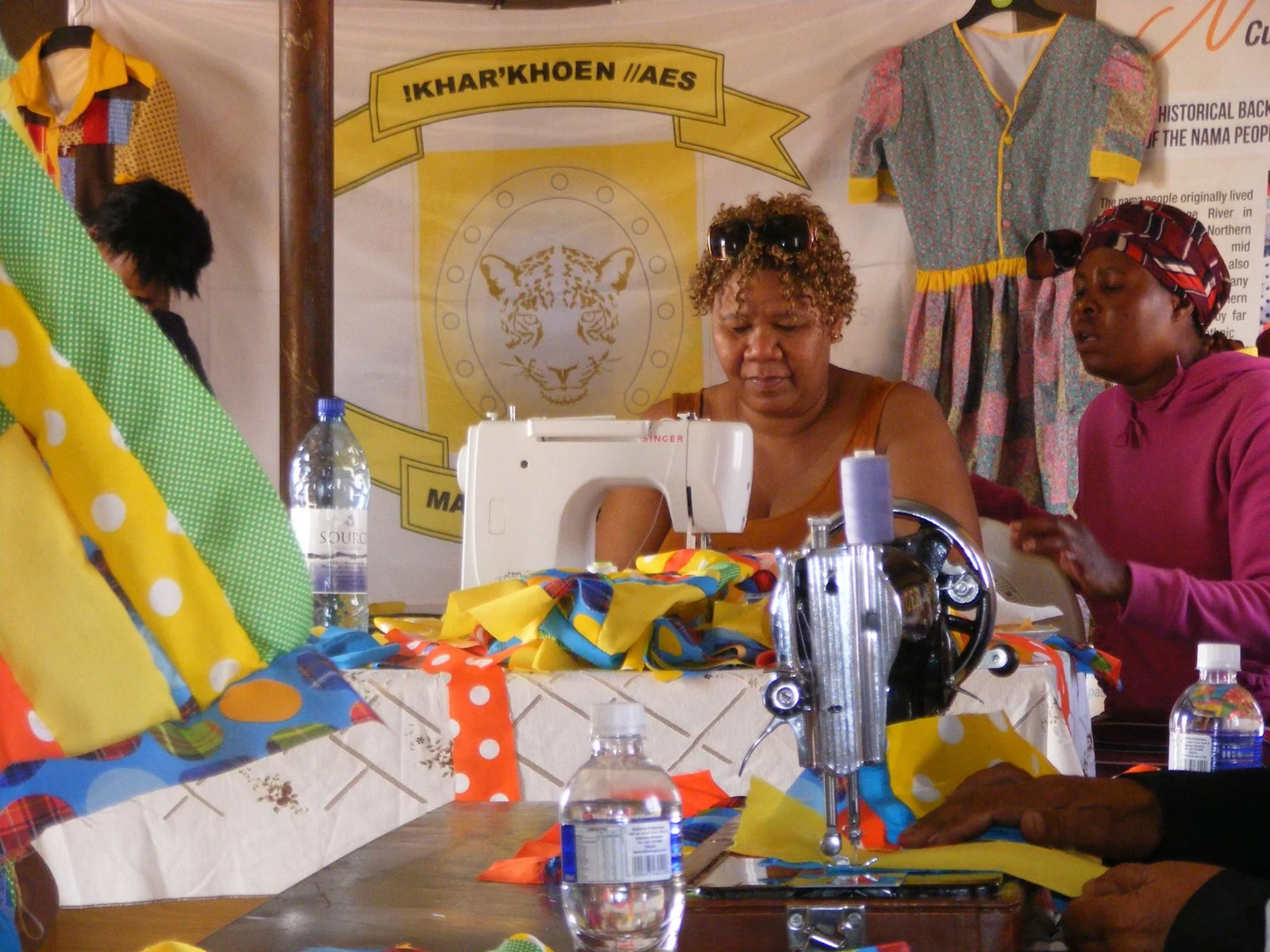 Women gather at sewing machines with colourful cloth and hanging garments during a patchwork workshop; the photographer identifies the event as Nama patchwork training in Lokgwabe.