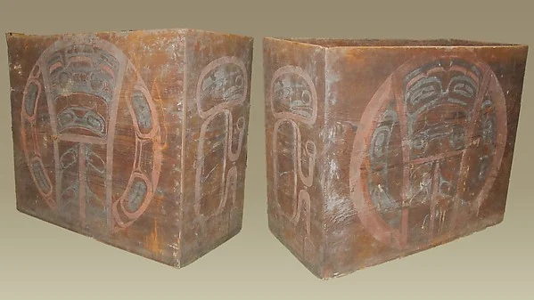 Two museum views of the same weathered wooden box drum, showing faded red and dark painted faces on its broad sides.