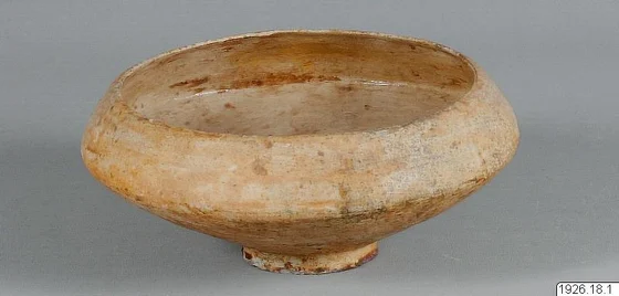 A shallow footed ceramic bowl with pale, reddish-brown-stained surfaces, photographed against grey with an inventory label.