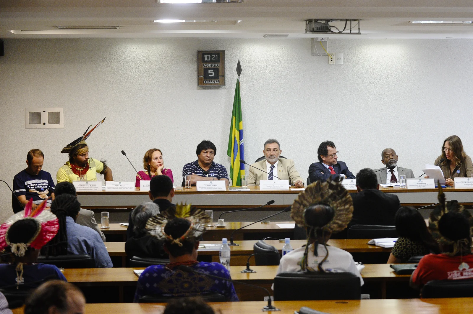 Kâhu Pataxó, wearing a yellow shirt and feather headdress, sits second from the left in a panel of eight speakers at a Brazilian Senate hearing, with audience members in the foreground.