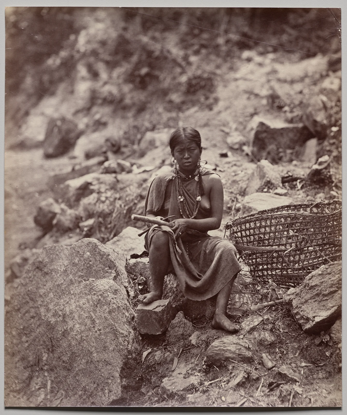 Historical sepia photograph of a seated woman on rocks, holding a cylindrical object beside a large openwork basket.