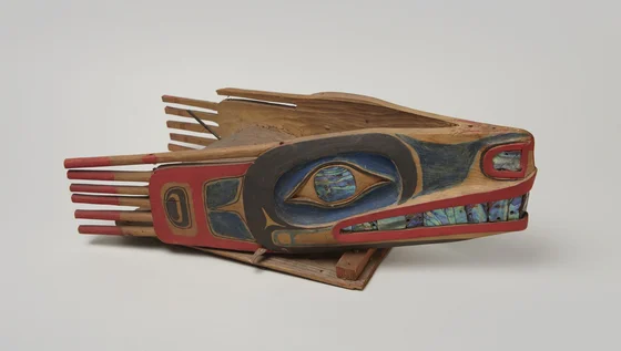 A carved wooden wolf-salmon trap headdress with a long muzzle, red and dark painted forms, abalone eye and teeth, and projecting wooden rods at the back.