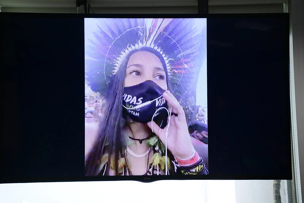 A Senate display shows Samela Sateré Mawé speaking remotely, wearing a dark face mask and feather headdress and holding an earbud microphone; the full monitor and surrounding room remain visible.