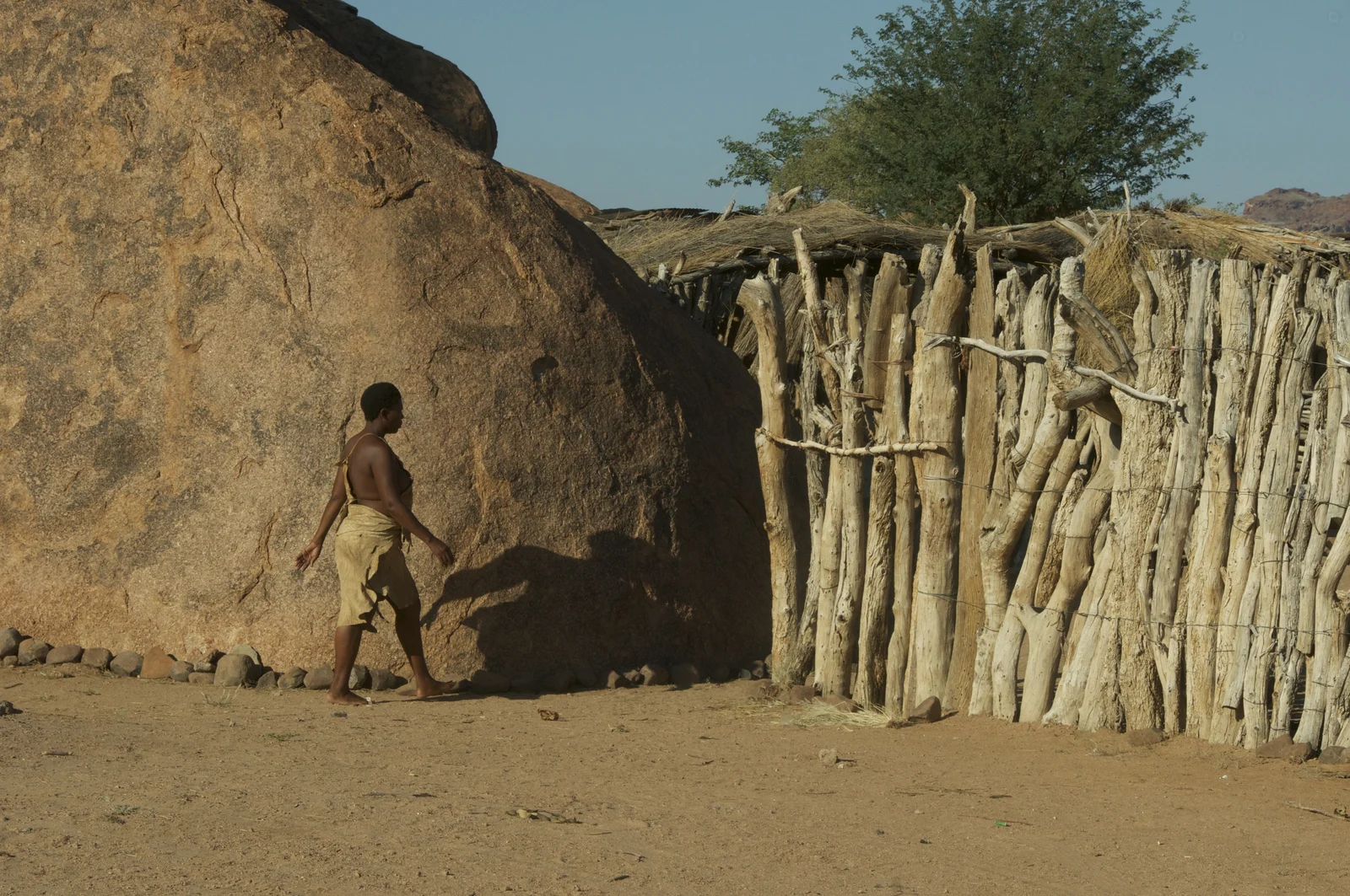 A person walks beside a large rock and a timber-and-thatch structure at the Damara Living Museum in Namibia.