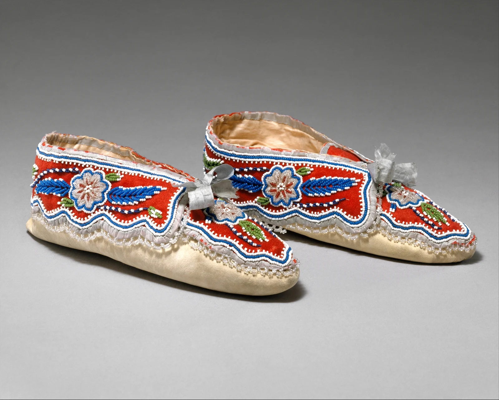 Pair of moccasins with red panels, blue and white floral beadwork, pale soles and silver-grey ribbons.