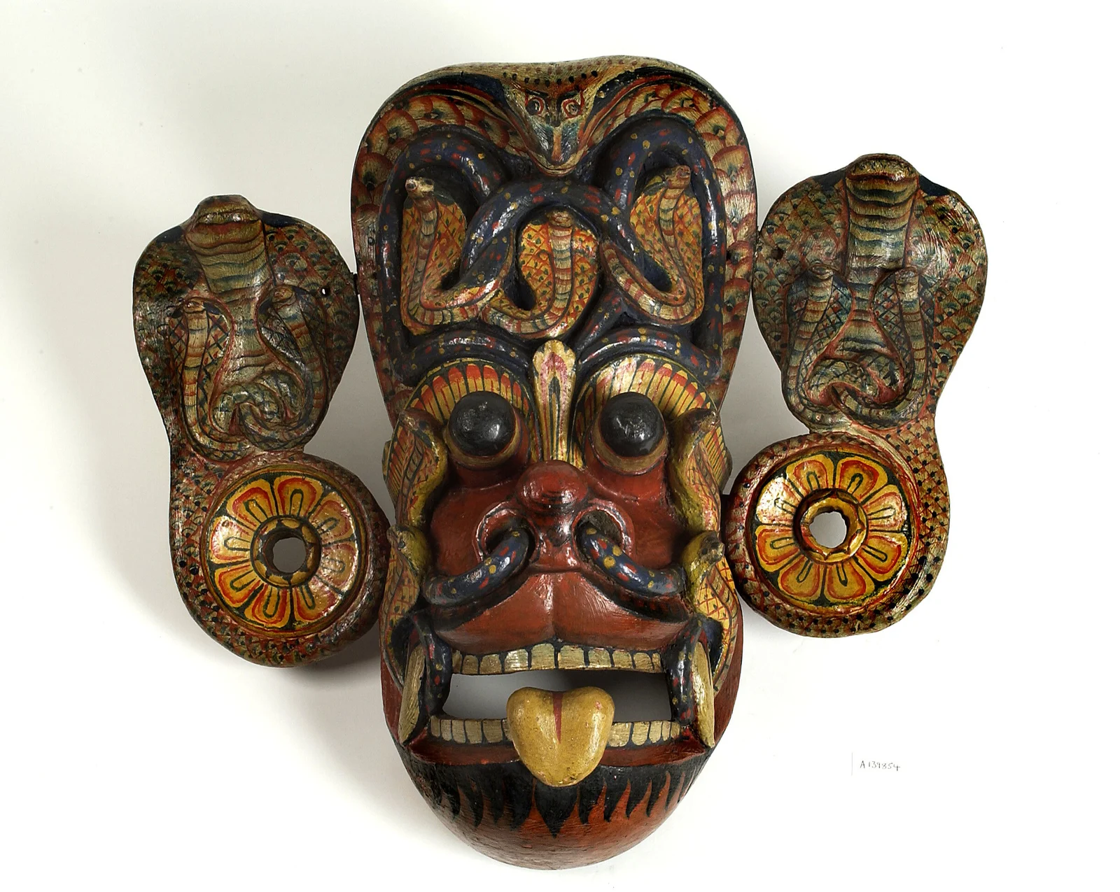 Worn painted wooden mask with a red face, bulging black eyes, exposed teeth and fangs, yellow protruding tongue, cobra canopy, serpents at the nostrils and separate cobra-and-lotus ear panels.