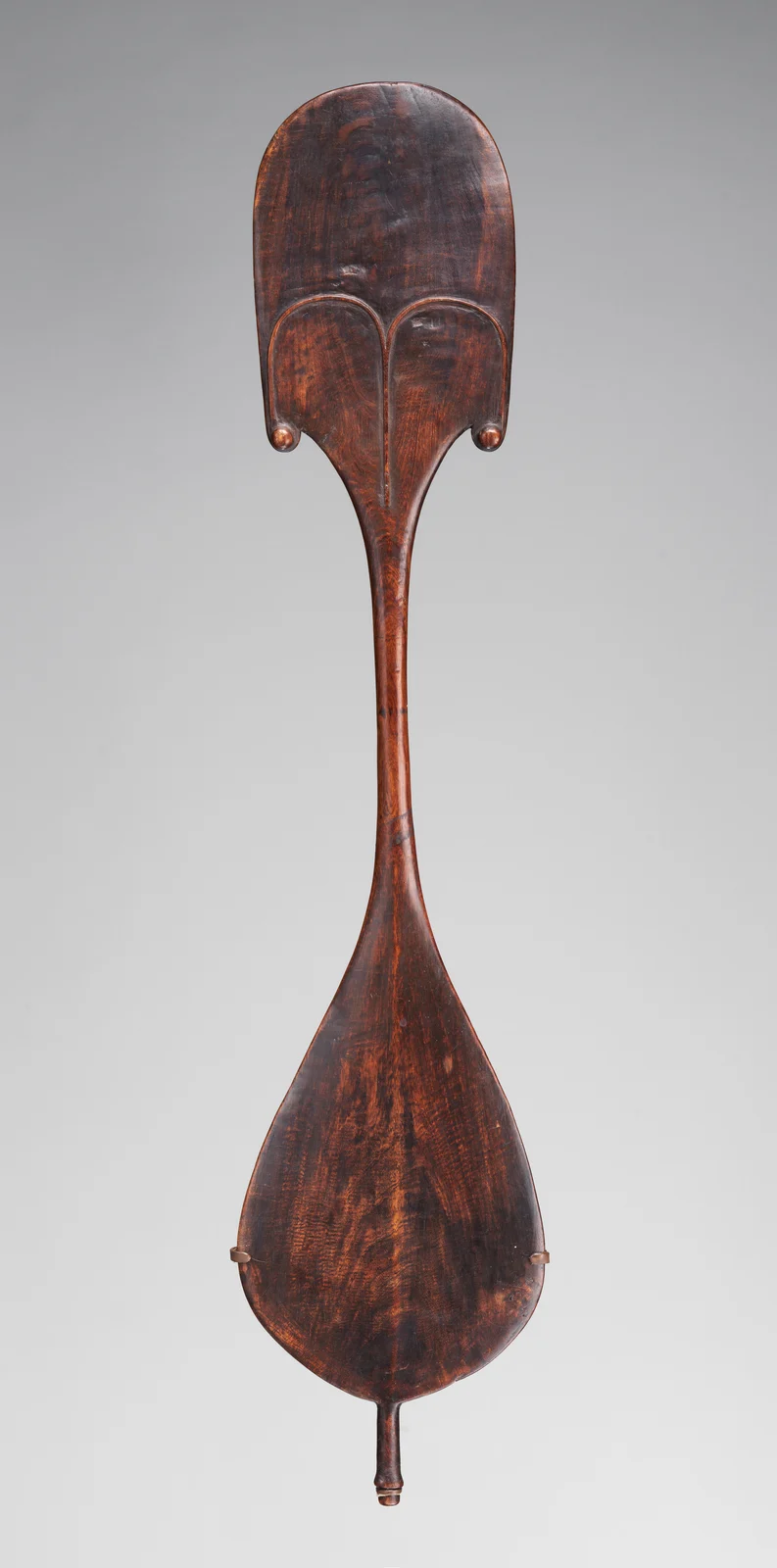 Dark wooden dance paddle with two broad lobes, a carved brow and nose on the upper lobe, and a narrow connecting shaft.