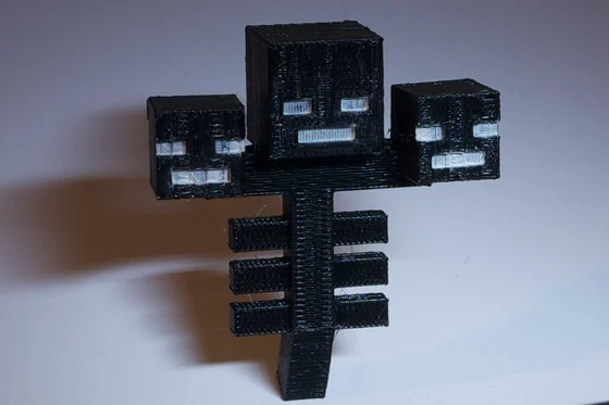 Black 3D-printed Minecraft Wither sculpture with three square skulls, white painted eyes and mouths, and a ribbed central body, photographed on a pale surface.