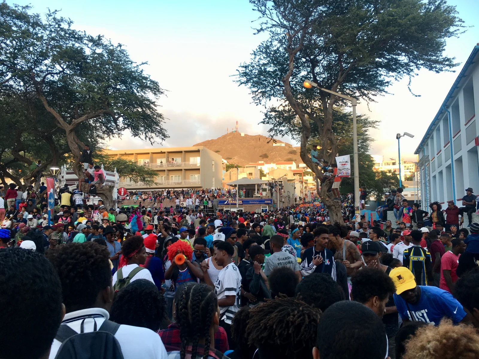 A dense carnival crowd fills a street and surrounding viewing areas beneath trees in Mindelo, with colourful costumes, urban buildings and a hill behind.