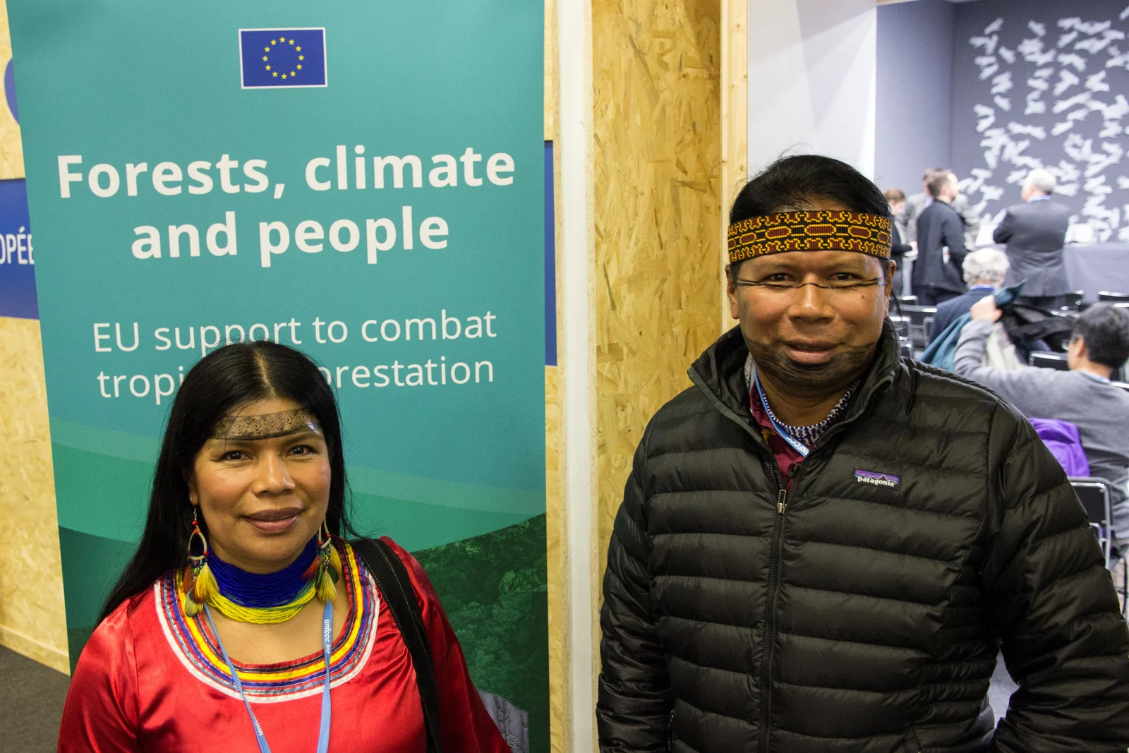 Patricia and José Gualinga stand beside a green Forests, climate and people display at COP21, with a meeting area visible behind them.