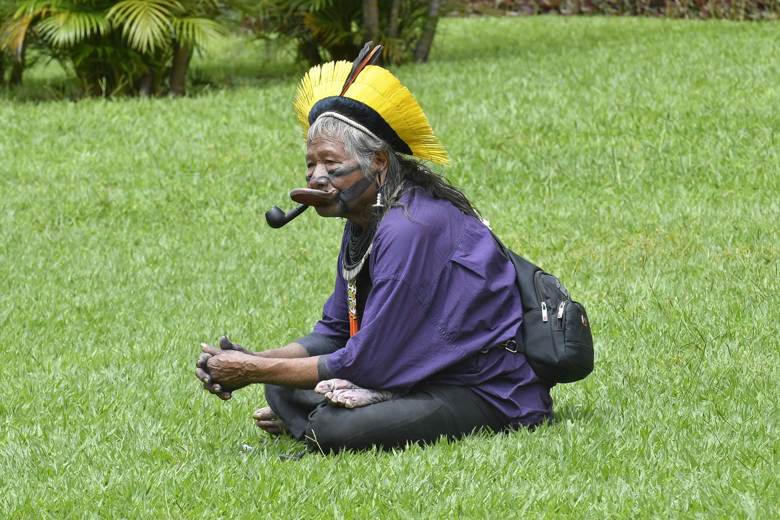 Raoni Metuktire sits cross-legged on grass during a2019 Senate visit, wearing a purple shirt and yellow feather headdress, with a black backpack and pipe.