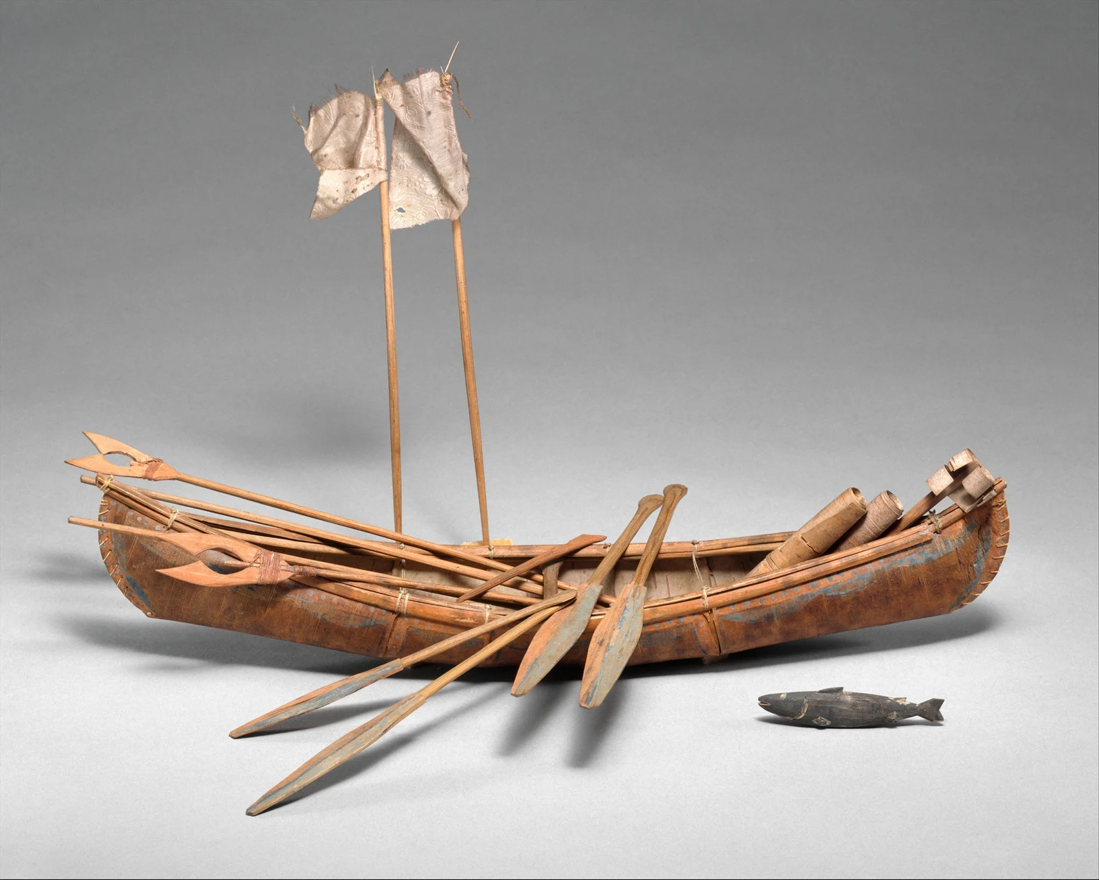A decorated miniature birchbark canoe with paddles, poles, small tools and two upright fabric flags, with a small fish-shaped object beside it.