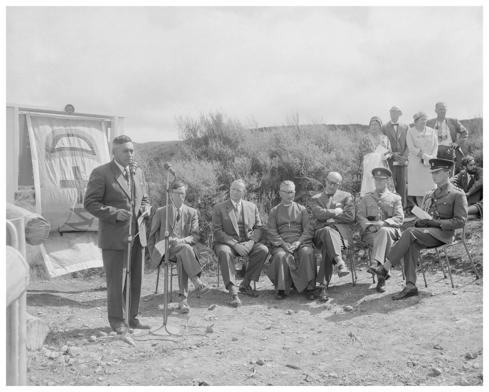 Hepi Te Heu Heu standing at microphones beside a covered commemorative plaque while officials and other attendees sit or stand outdoors, 18 February 1961.