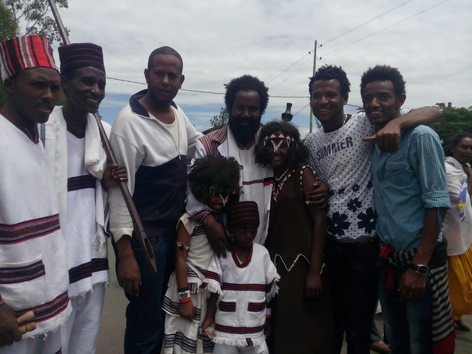 A group poses outdoors in a mixture of striped white garments, bead adornments, caps and everyday shirts; the photographer identifies the occasion as an Oromo Irreecha event.