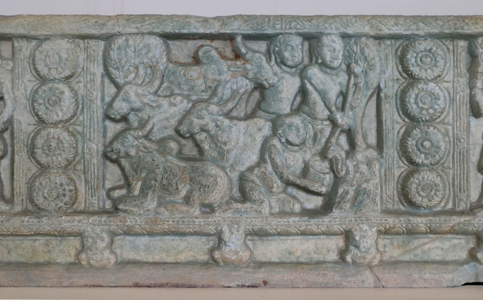 Weathered stone relief with cattle at left and human figures beneath trees at right, one crouching beside a vessel and the small serpent in the capture scene.