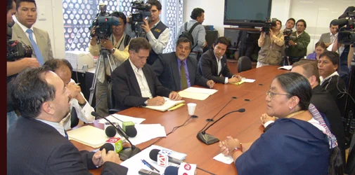 Nina Pacari sits at the near right of a conference table facing officials, with microphones, documents and press cameras around the room.
