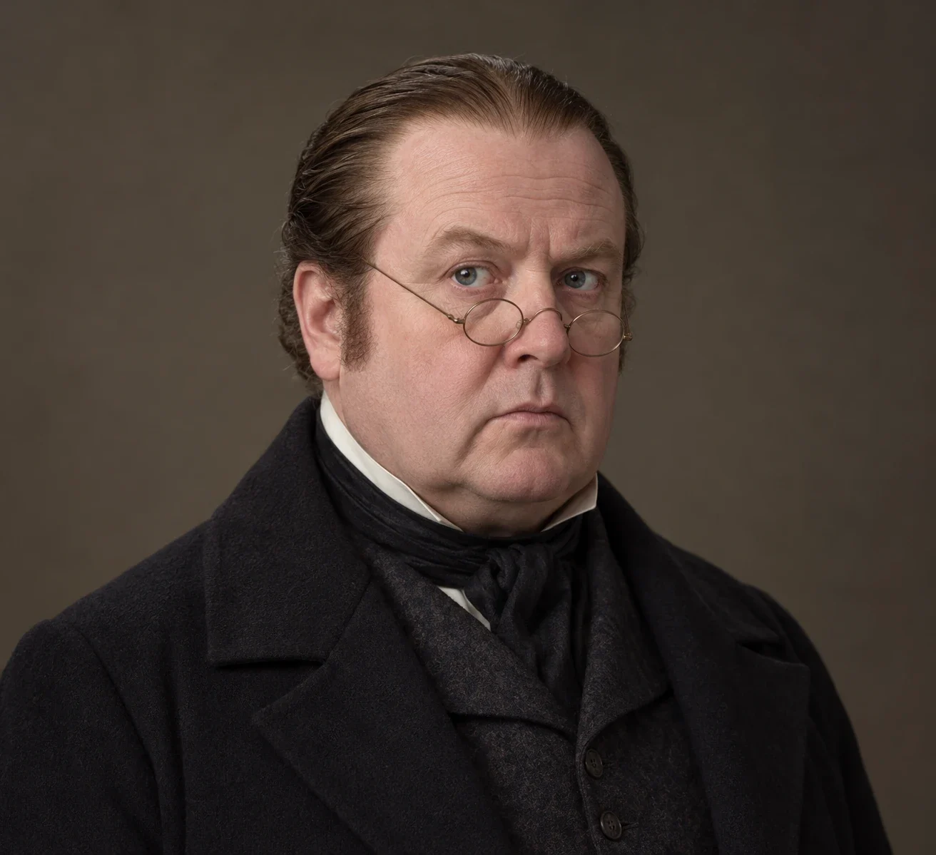 Drumknott in the 2010 television interpretation: pale round face, slicked-back brown hair, small round spectacles low on the nose, dark formal coat, pale collar and dark neckcloth.