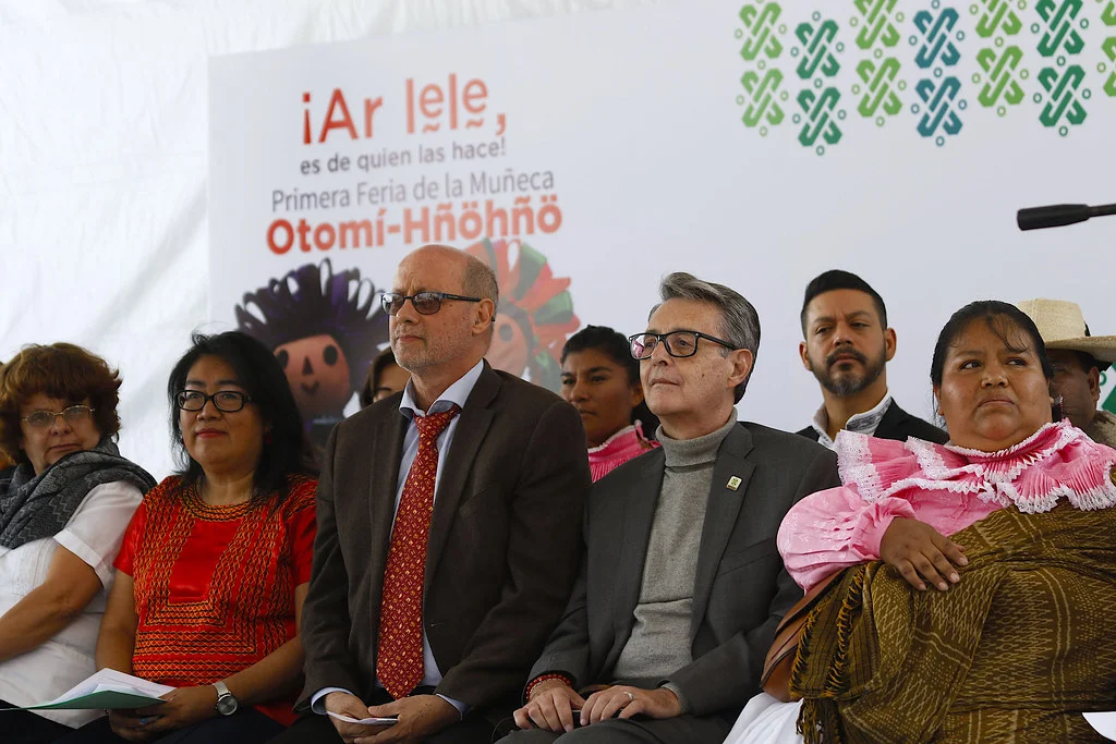 Seated attendees at the opening of the first Ar lele Otomí-Hñöhñö Doll Fair, in front of a banner naming the fair and showing a doll illustration.