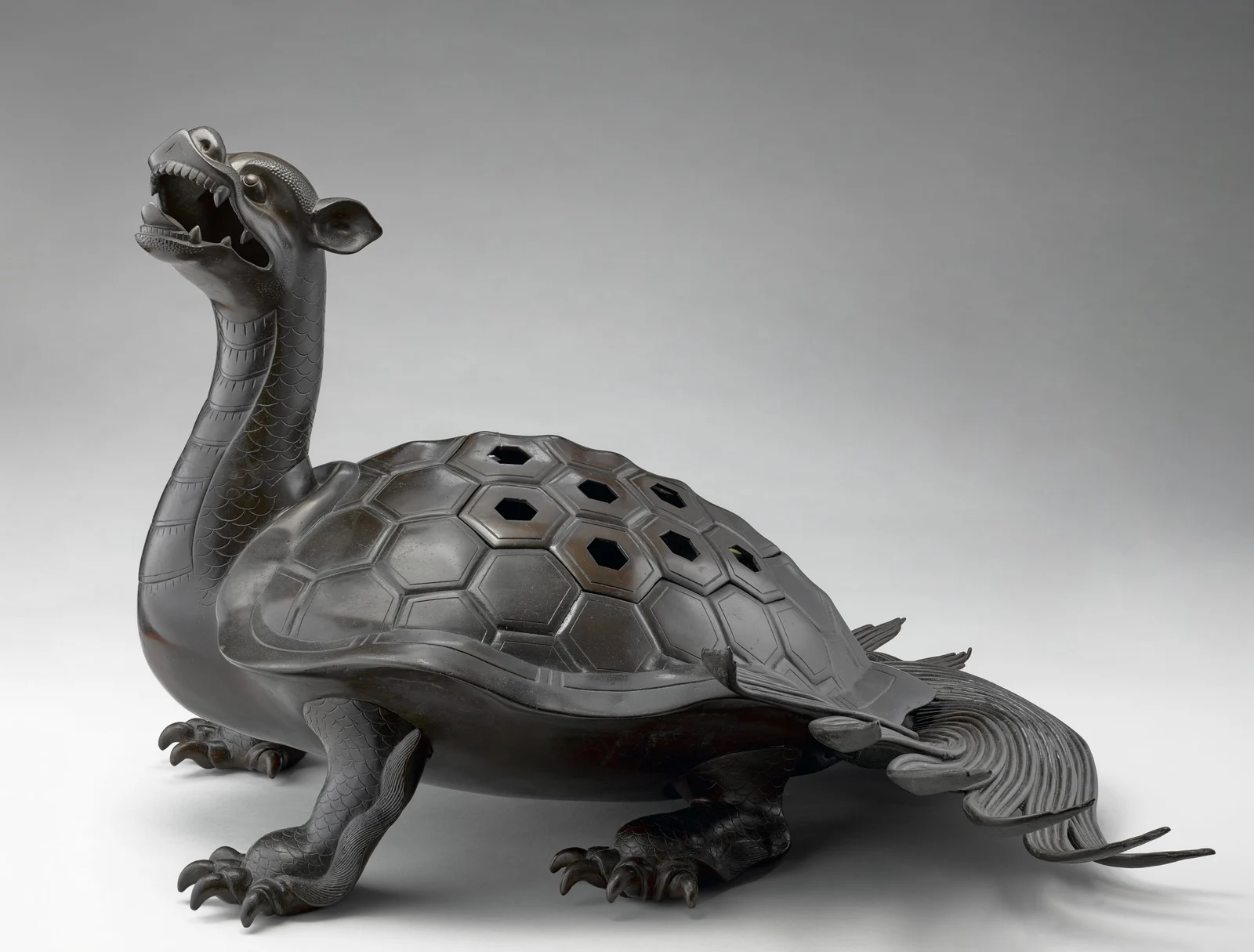 Dark bronze tortoise incense burner with a high upright neck, open toothed mouth, pointed ears, clawed feet, perforated hexagonal shell and a long curling tail shaped as flowing strands of algae.