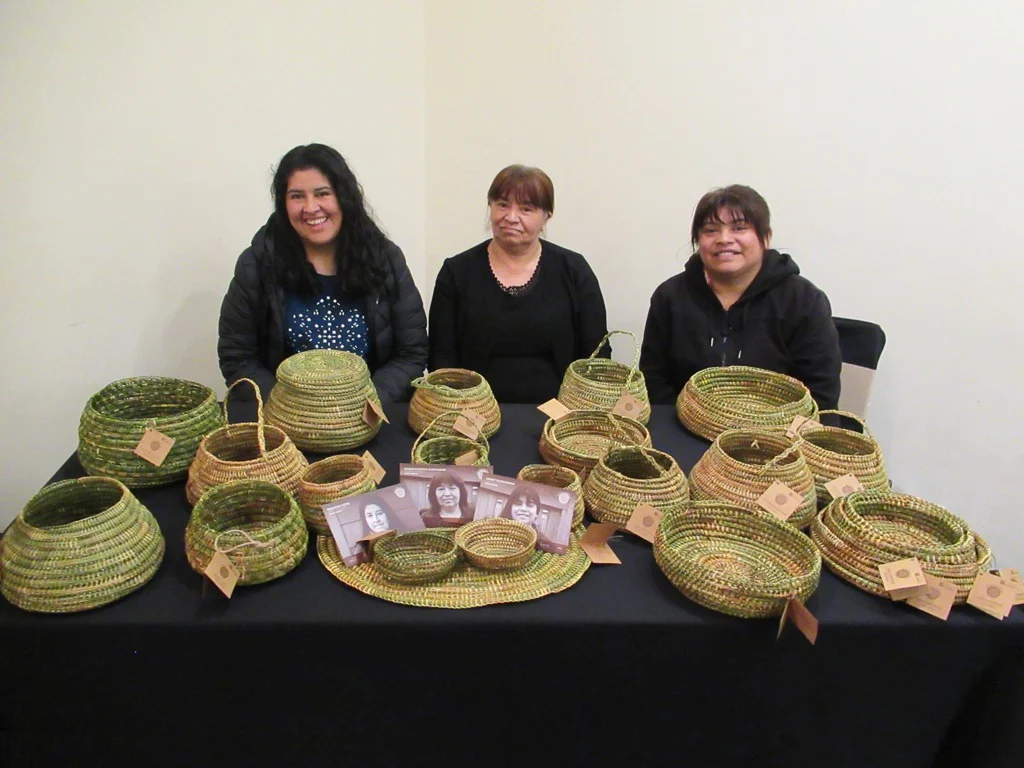 Three basket makers sit behind a table displaying rounded woven baskets, handled baskets and shallow trays, with small portrait cards among the work.