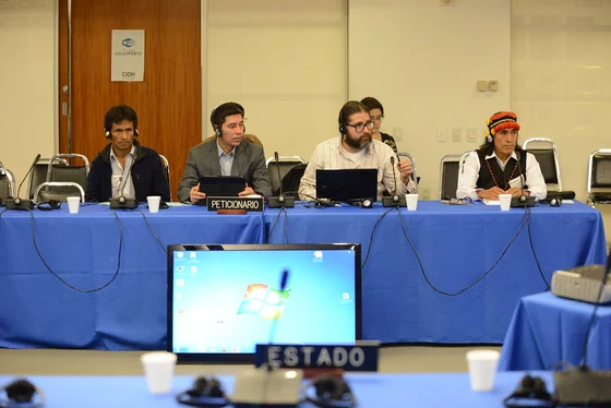 Four petitioners sit behind a blue-covered hearing table with headsets and microphones; Domingo Ankuash of Federación Shuar is seated at the right.